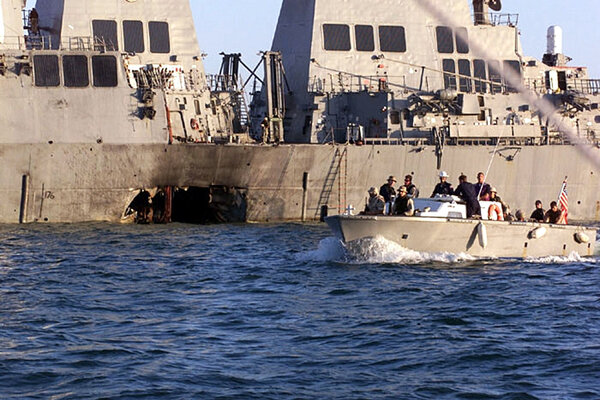 A small boat sails past a large military ship with a hole in its side.