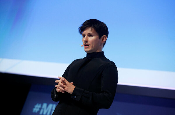 Pavel Durov, wearing a dark shirt, speaks while standing before a blue screen.