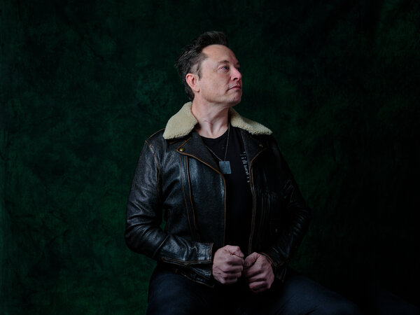 Elon Musk wears a dark leather jacket with a fur collar against a dark background.